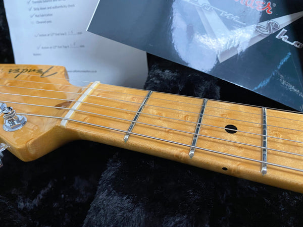 Close-up view of Fender guitar maple neck and fretboard with metal frets and strings, showing birdseye maple wood grain on black plush background with Fender certificate and papers nearby.