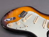 Close-up of a sunburst electric guitar body with white pickguard, three single-coil pickups, chrome bridge, and tone and volume control knobs on grey background
