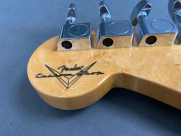 Close-up of Fender Custom Shop logo and chrome tuning pegs on natural wood guitar headstock