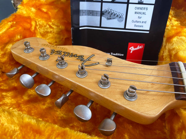 Close-up of Fender Telecaster guitar headstock with tuning pegs, set against a bright orange plush guitar case interior and a Fender owner's manual in the background.