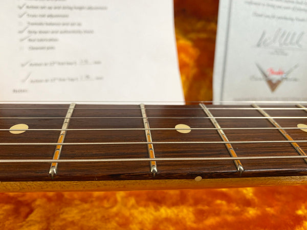 Close-up of guitar fretboard with dot inlays and steel strings on a vintage acoustic guitar with certificate and checklist blurred in background.