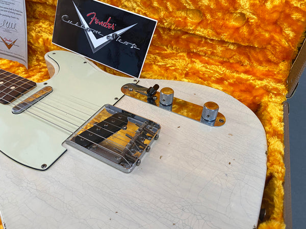 Close-up of vintage white Fender Telecaster electric guitar with visible wear and cracking on the body, chrome bridge, control knobs, and a white pickguard, displayed in an orange plush-lined guitar case with Fender Custom Shop certificate.