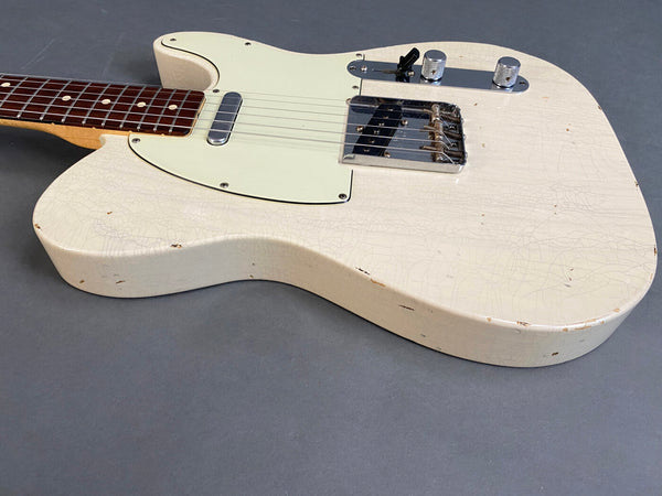Close-up of white vintage-style electric guitar with worn cracked finish, cream pickguard, single-coil pickups, and rosewood fretboard on gray background