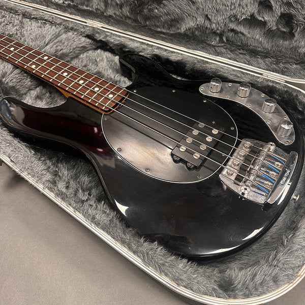 Black electric bass guitar with rosewood fretboard, chrome bridge and control knobs, resting in a gray plush-lined hard case