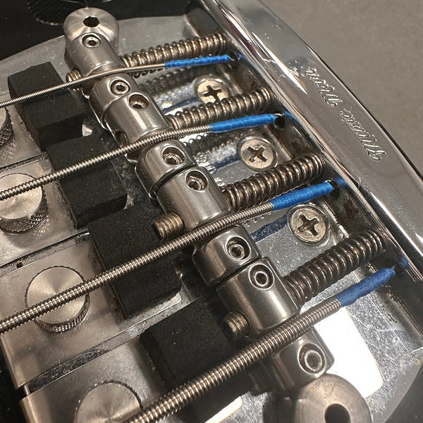 Close-up of electric bass guitar bridge with metal saddles, springs, and steel strings with blue thread windings at Coffee House Guitars