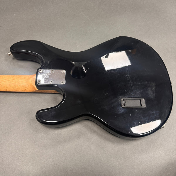 Black electric guitar body back with scratches and a maple neck joint on gray background, showing metal neck plate and battery compartment cover