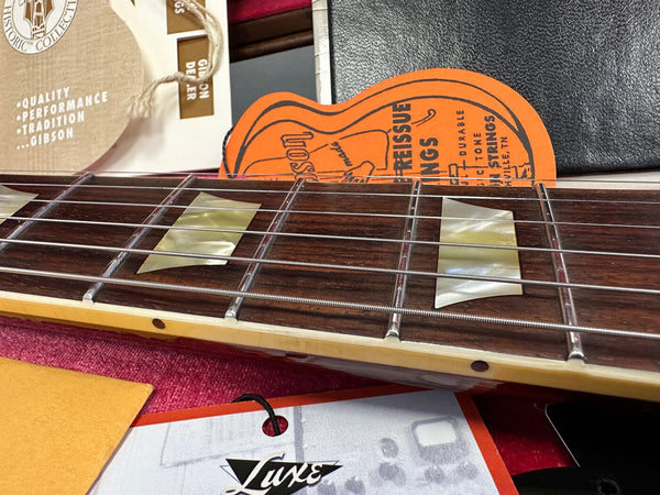 Close-up of Gibson guitar rosewood fretboard with trapezoid inlays and metal strings showing detailed craftsmanship and vintage Gibson paper tags in background