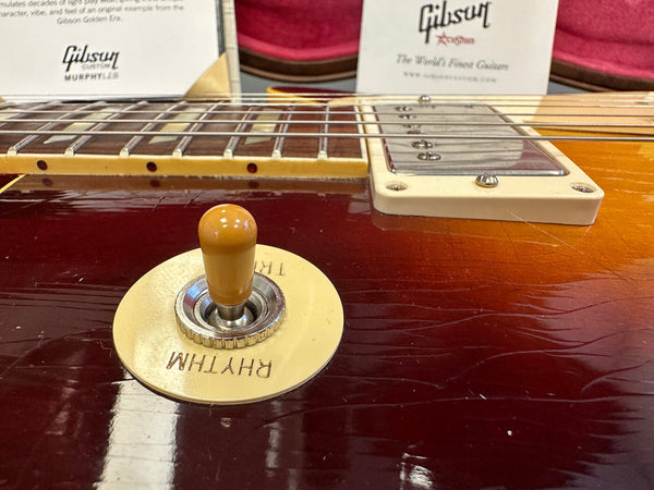 Close-up of vintage Gibson Les Paul guitar toggle switch labeled "Rhythm" and "Treble" with aged sunburst finish and cream pickup mounting ring, featuring Gibson Custom Murphy Lab and Gibson USA branding in background.