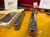 Close-up of Gibson electric guitar bridge and tailpiece on a sunburst finish body with chrome hardware and amber control knobs, featuring Gibson product information card in the background