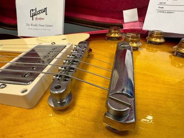 Close-up of Gibson electric guitar bridge and tailpiece on a sunburst finish body with chrome hardware and amber control knobs, featuring Gibson product information card in the background