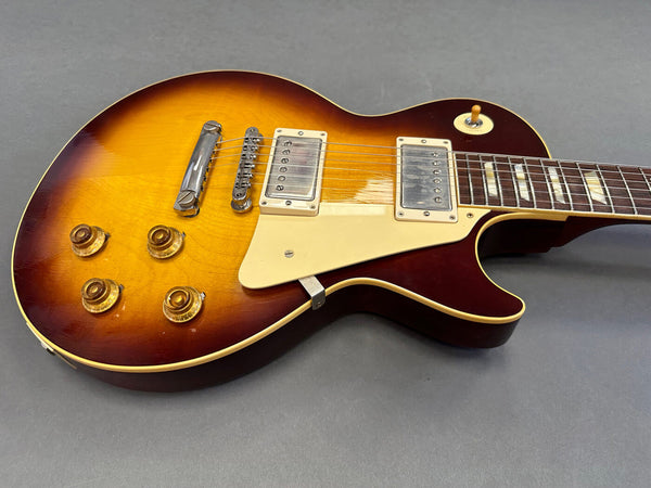 Close-up image of a sunburst electric guitar with dual humbucker pickups, four control knobs, and a cream-colored pickguard on a grey surface.