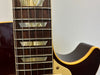 Close-up of electric guitar fretboard with trapezoid inlays, metal frets, and dark wood grain, showing part of dark brown body with cream binding and pickguard.
