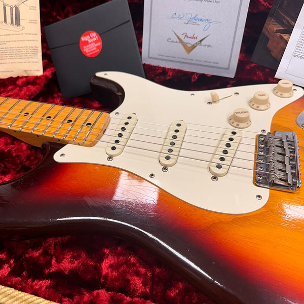 Close-up of vintage sunburst Fender Stratocaster electric guitar showing maple neck, white pickguard, three single-coil pickups, tone and volume knobs, and worn finish on red plush guitar case lining with Fender certification and accessory box in background.