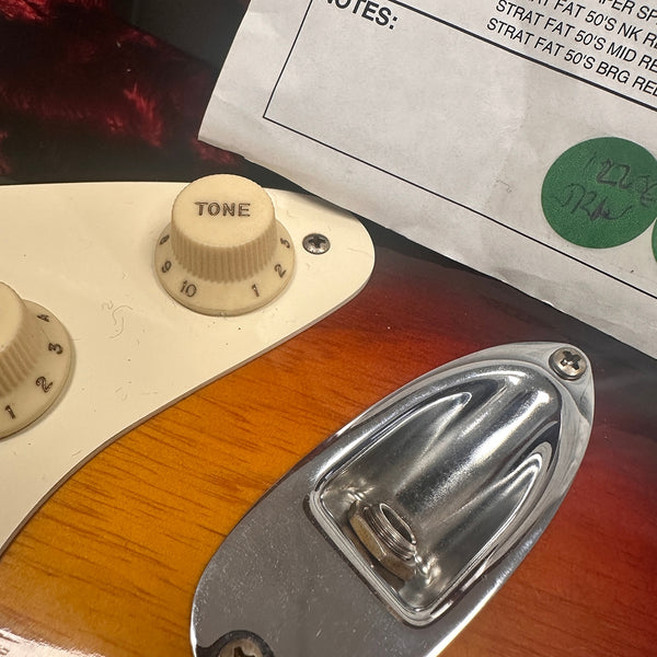 Close-up of sunburst electric guitar body showing cream tone control knob and chrome output jack plate with paper listing guitar pickup details above