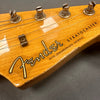 Close-up of Fender Stratocaster guitar headstock with vintage tuning pegs and strings, natural wood finish, showing Fender logo and "with synchronized tremolo" text, with visible wear on the edge.