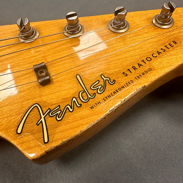 Close-up of Fender Stratocaster guitar headstock with vintage tuning pegs and strings, natural wood finish, showing Fender logo and "with synchronized tremolo" text, with visible wear on the edge.