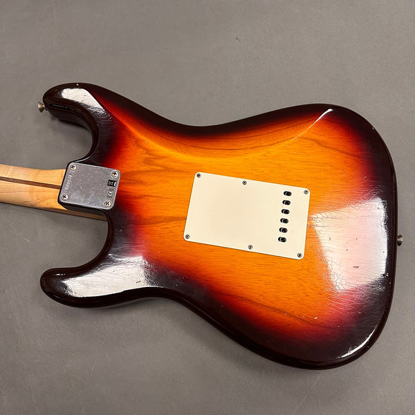 Vintage sunburst electric guitar body with a white backplate and visible wear and aging on the finish and wood grain, viewed from the back on a gray surface.