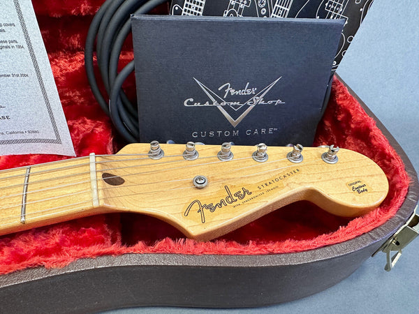 Close-up of Fender Stratocaster guitar headstock in vintage hard case with red plush interior, showing tuning pegs and Fender Custom Shop certificate in background.