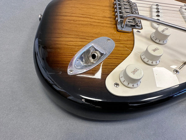 Close-up of sunburst electric guitar body showing input jack, tone and volume control knobs, white pickguard, and tremolo arm on gray background