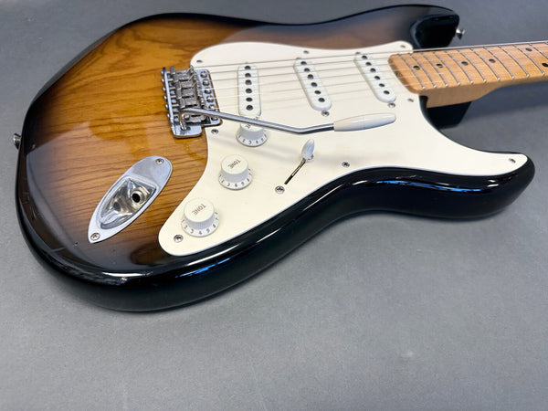 Close-up of sunburst electric guitar body with white pickguard, three single-coil pickups, tone and volume control knobs, and tremolo arm on gray background