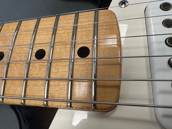 Close-up of electric guitar maple fretboard with metal frets and black dot inlays, showing steel strings and part of white guitar body.