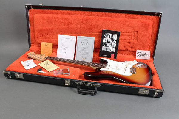 Fender Stratocaster electric guitar in sunburst finish inside black hard case with orange plush interior, including Fender manuals and accessories.