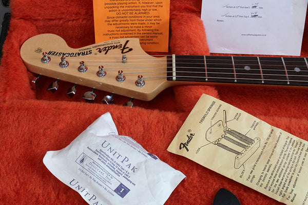 Fender Stratocaster guitar headstock with tuning pegs, rosewood fretboard, and orange guitar case interior, accompanied by Fender tremolo springs instruction sheet, humidifier unit pak, and setup adjustment notes from Coffee House Guitars.