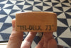 Close-up of wooden guitar part labeled "TEL-DELX. 73" held by hand over geometric patterned floor background