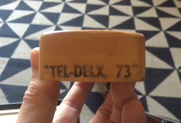 Close-up of wooden guitar part labeled "TEL-DELX. 73" held by hand over geometric patterned floor background
