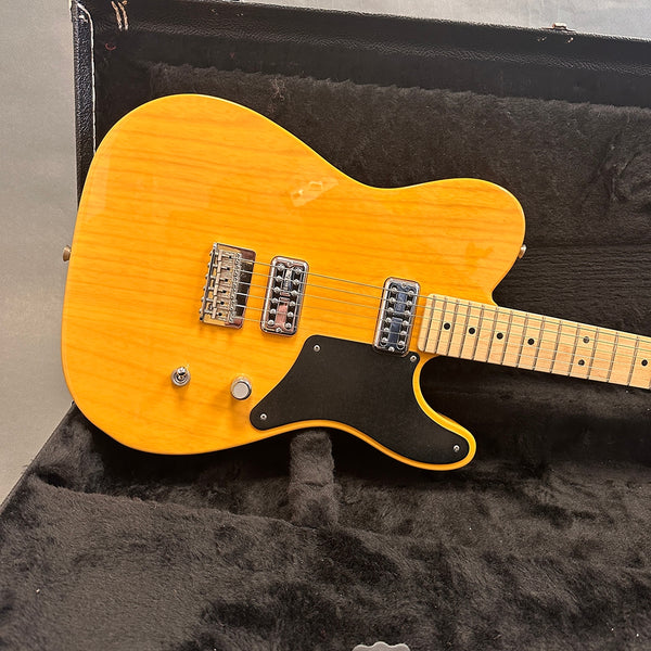 Electric guitar with natural wood finish, black pickguard, dual chrome pickups, and maple neck, resting in a black plush guitar case interior.