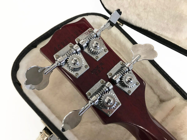 Close-up of bass guitar headstock with chrome Grover tuning pegs and dark wood finish inside plush guitar case