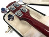 Close-up view of red wood bass guitar headstock with chrome tuning pegs resting in plush white guitar case interior