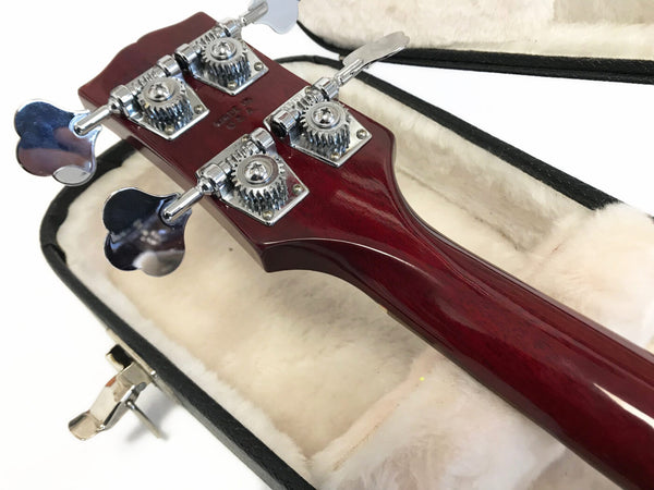 Close-up view of red wood bass guitar headstock with chrome tuning pegs resting in plush white guitar case interior
