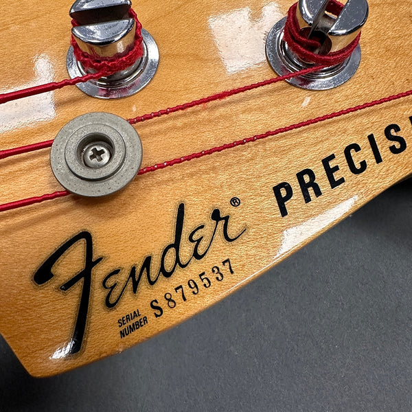 Close-up of Fender Precision Bass guitar headstock showing tuning pegs, red strings, and serial number S879537 on natural wood finish