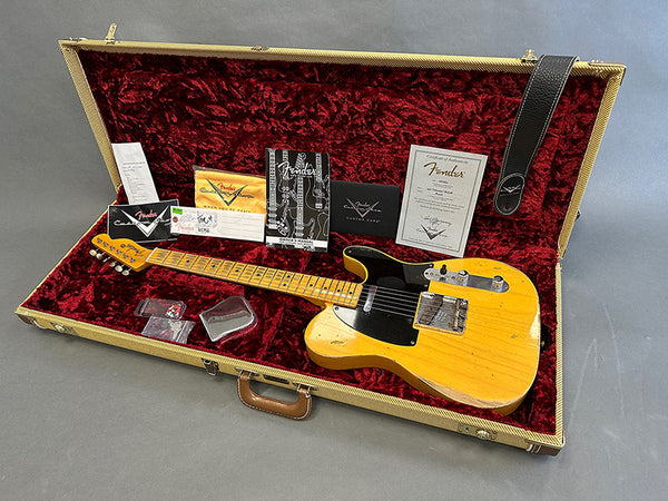 Fender vintage-style electric guitar with maple neck and black pickguard in tweed hard case with red velvet interior, including Fender manuals, certificates, strap, and accessories