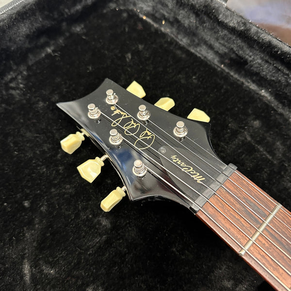 Close-up of black electric guitar headstock with cream tuning pegs, rosewood fretboard, and "McCarthy" logo on a black plush case interior.