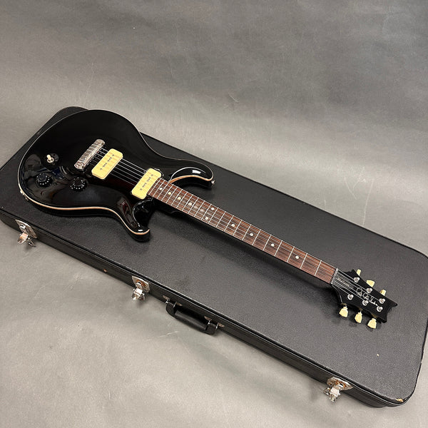 Black electric guitar with cream pickups and knobs on black hard case, six-string with rosewood fretboard and dot inlays, guitar headstock with tuning pegs and logo, studio background.