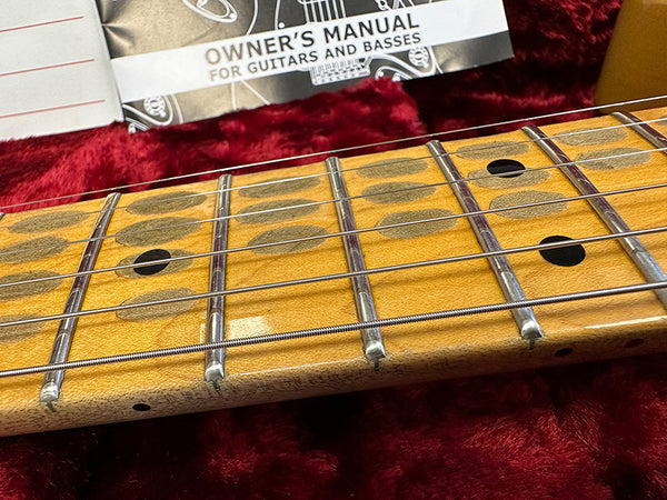 Close-up of maple guitar fretboard with metal frets and six strings, resting on red plush interior of guitar case with owner's manual for guitars and basses in the background