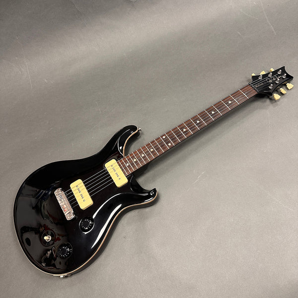 Black electric guitar with dual cream-colored pickups, rosewood fretboard, and chrome hardware on grey background