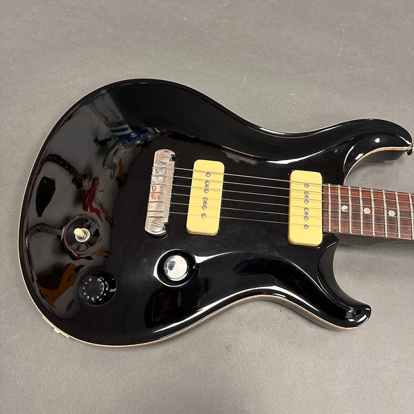Close-up of black electric guitar body with cream-colored P90 pickups, chrome bridge, black tone knob, and toggle switch on grey background.