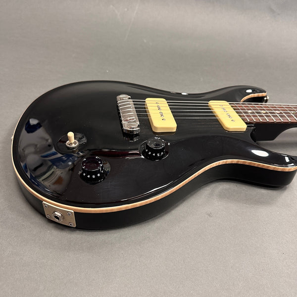 Black electric guitar with cream-colored pickups, black control knobs, and cream binding on body edges, shown on gray background.