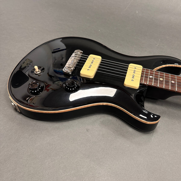 Close-up of black electric guitar body with cream-colored P90 pickups, chrome bridge, black control knobs, and toggle switch on grey background