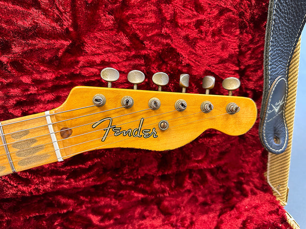 Close-up of Fender electric guitar headstock with vintage tuning pegs and logo on maple neck, resting on red plush guitar case lining with Fender strap visible