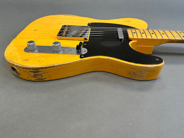 Close-up of vintage yellow electric guitar body with black pickguard and chrome control knobs showing worn finish and wood grain texture on gray background.