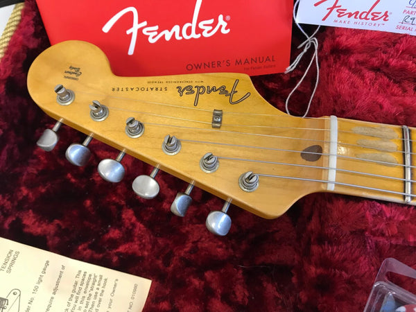 Close-up of Fender Stratocaster guitar headstock with tuning pegs and string trees on red plush case lining, showing Fender owner's manual in background.