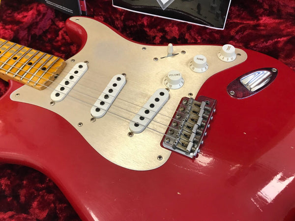 Close-up of red electric guitar body with three single-coil pickups, white pickguard, volume and tone control knobs, and vintage-style bridge on red plush background