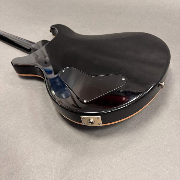 Back side of a black electric guitar with a glossy finish, featuring a control cavity cover and metal output jack plate, photographed on a gray surface.