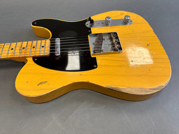 Vintage blonde Telecaster electric guitar with black pickguard and worn finish on body edges and lower bout, showing relic wear and aging signs, close-up angle on body and neck.