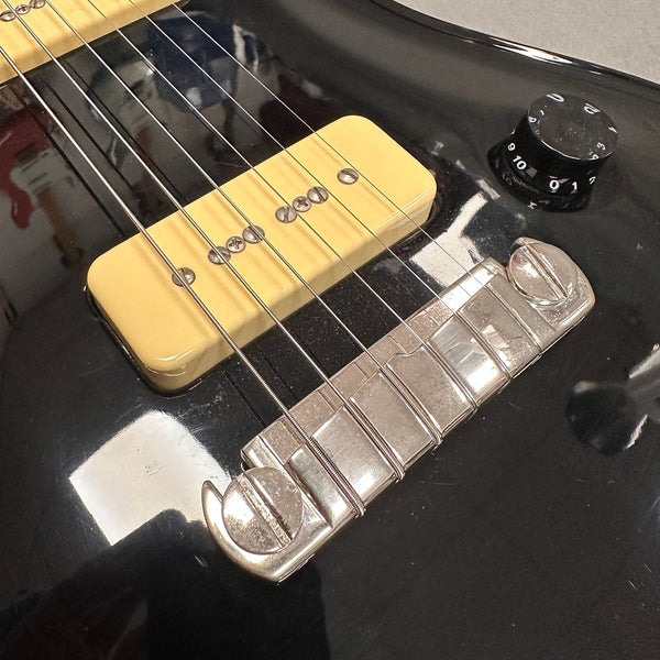 Close-up of black electric guitar body showing cream P90 pickup, metal bridge, strings, and black volume knob with white numbers and markings