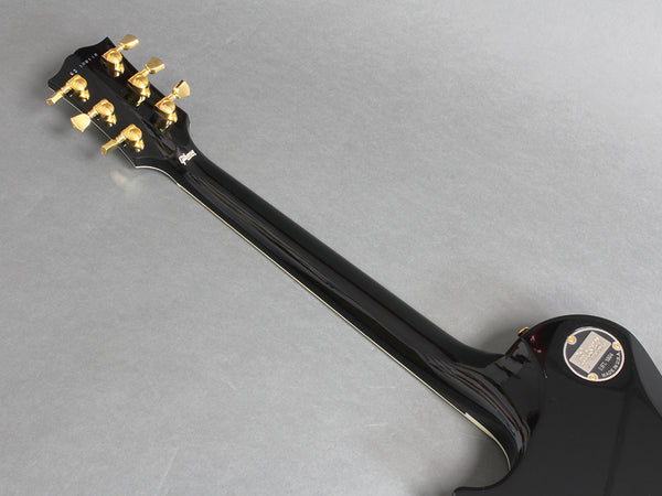 Back of black Gibson electric guitar neck showing gold tuning pegs and Gibson logo plate on a gray background